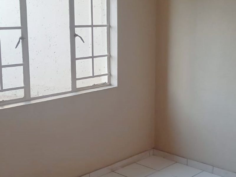 A clean, empty bedroom featuring white tiled flooring, neutral walls, and a window fitted with security bars.
