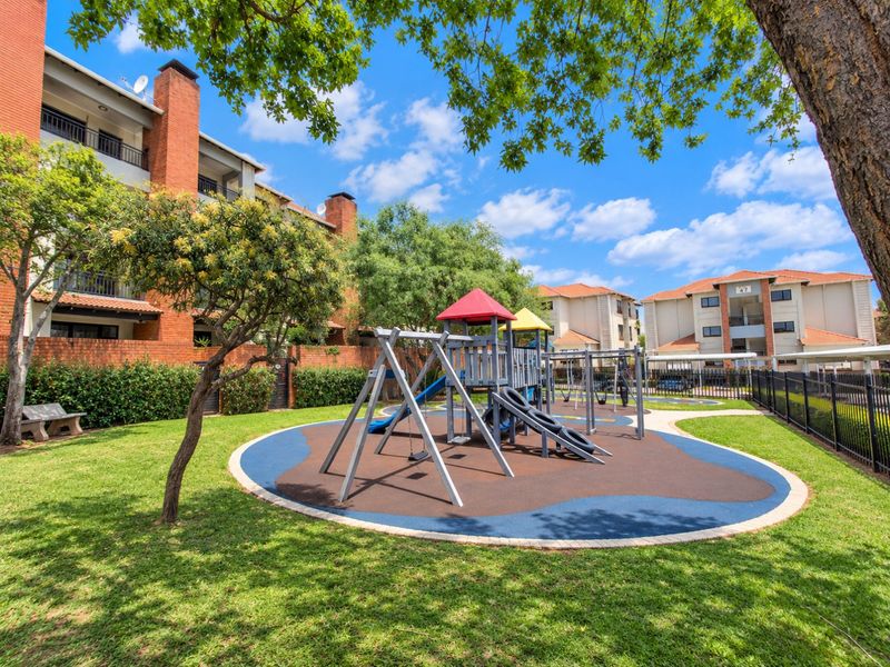 A well-maintained communal garden area featuring a modern playground with rubberized flooring, surrounded by landscaped lawns and apartment building facades.