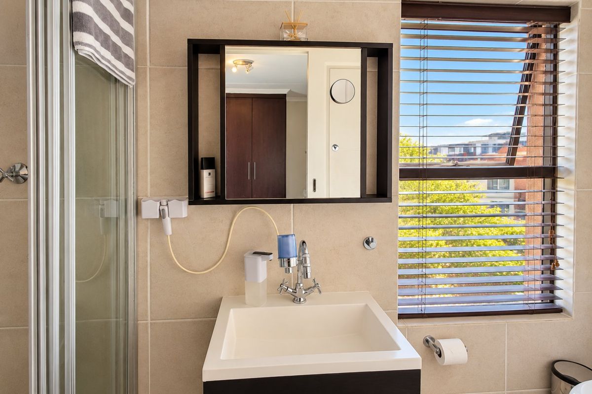 A well-maintained bathroom featuring a modern vanity basin, wall-mounted mirror cabinet, shower enclosure, and large window with venetian blinds.