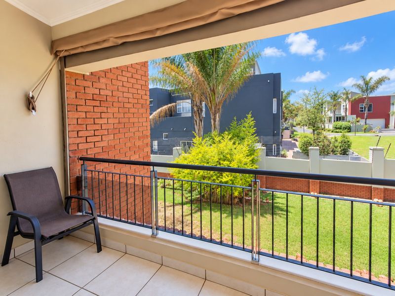 A covered balcony featuring tiled flooring, a brick accent wall, and a metal balustrade, offering a pleasant view of the surrounding garden and neighborhood.