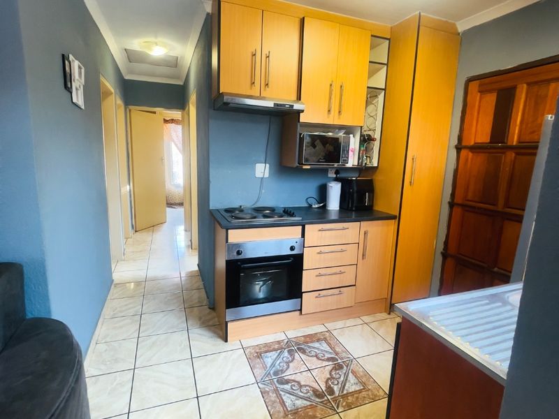 Compact kitchen featuring light wood cabinetry, built-in oven and hob, extractor fan, and tiled flooring, leading into a hallway.