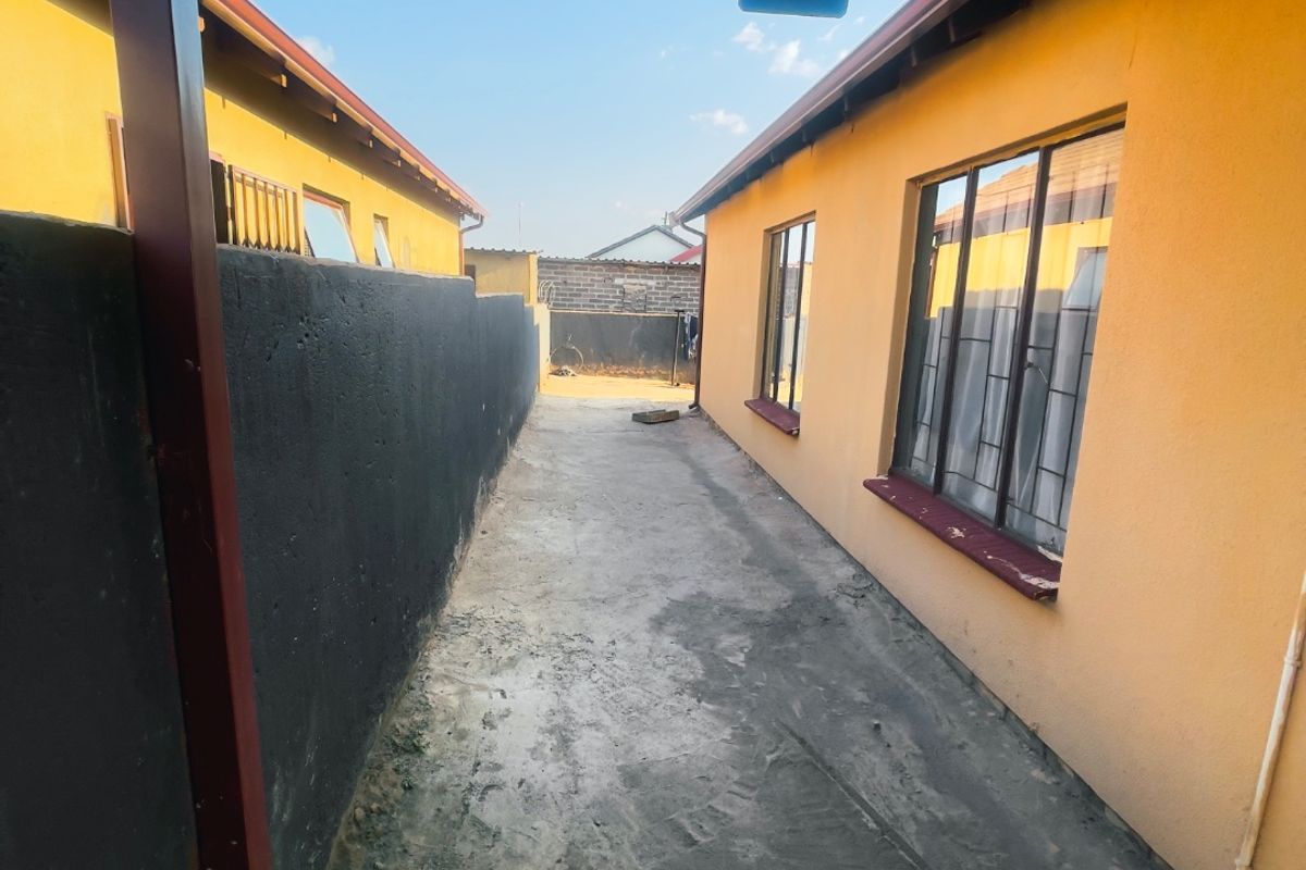 A narrow side passage between two yellow-painted structures, featuring a covered walkway with a corrugated metal roof, concrete paving, and security lighting.
