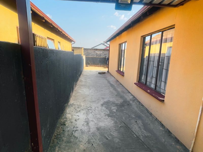 A narrow side passage between two yellow-painted structures, featuring a covered walkway with a corrugated metal roof, concrete paving, and security lighting.
