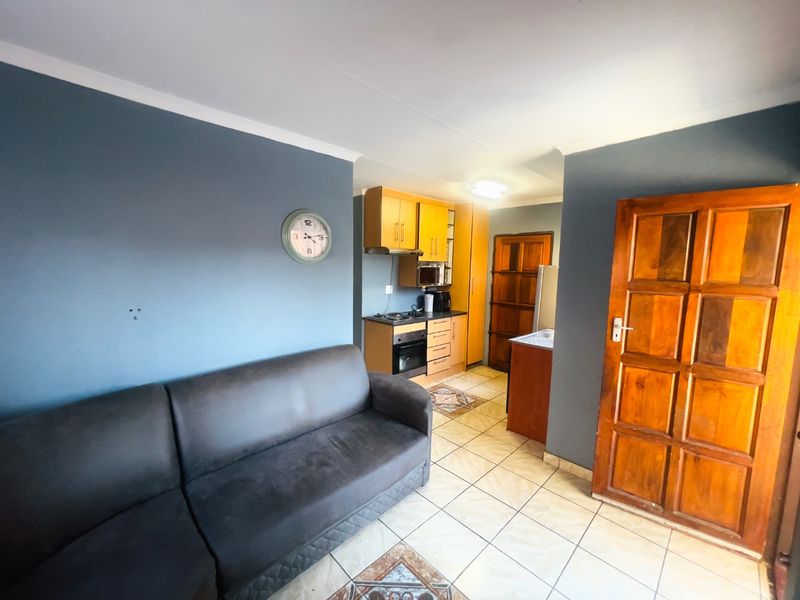 Open plan living area featuring a compact kitchen with built-in cabinetry, oven, and hob, finished with tiled flooring and a wooden entrance door.