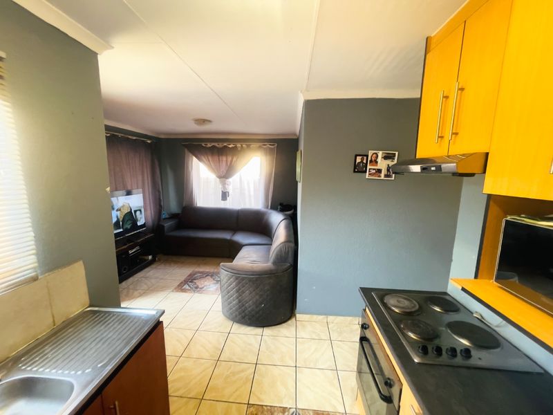 Open-plan living area featuring a functional kitchen with built-in cabinetry, electric hob, oven, and tiled flooring, seamlessly flowing into the adjacent lounge space.