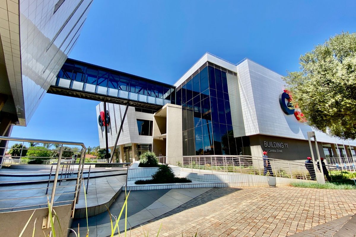 Modern commercial office building exterior featuring a glass curtain wall, elevated pedestrian bridge, and paved entrance area with professional landscaping.