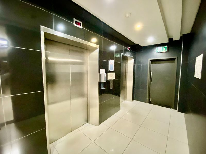 A modern commercial hallway featuring stainless steel elevator access, black tiled wall finishes, and a fire exit door with bright recessed lighting.