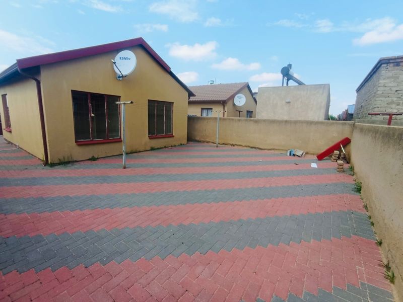 A rear exterior view of the house featuring a paved courtyard, boundary wall, and solar water heating system.