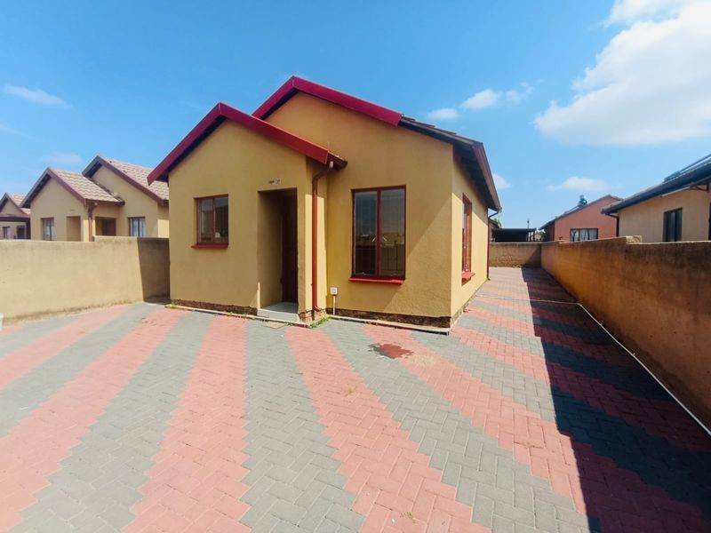 A detached single-story house featuring a paved driveway, perimeter boundary walls, and a pitched metal roof in a residential setting.