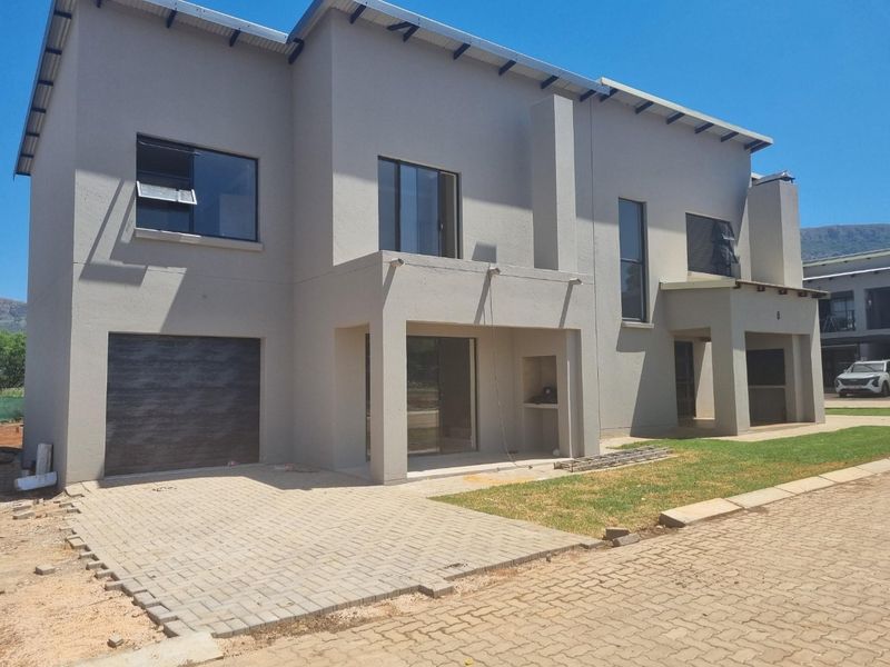 Modern double-storey townhouse exterior featuring a paved driveway, integrated garage, and a covered patio area with a built-in braai.