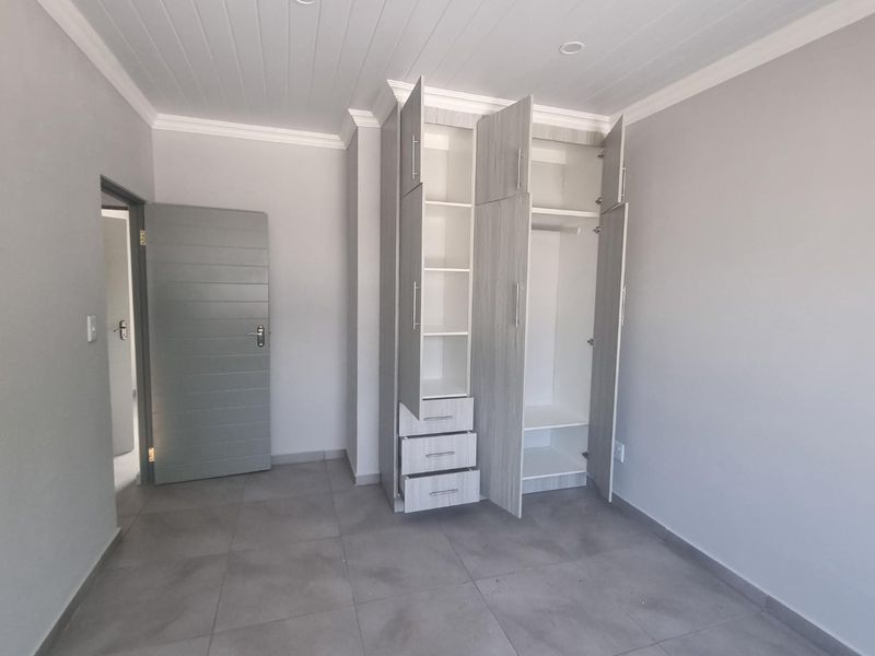 A clean, empty bedroom featuring modern grey tiled flooring, neutral walls, recessed ceiling lights, and functional built-in cupboards with integrated shelving and drawers.