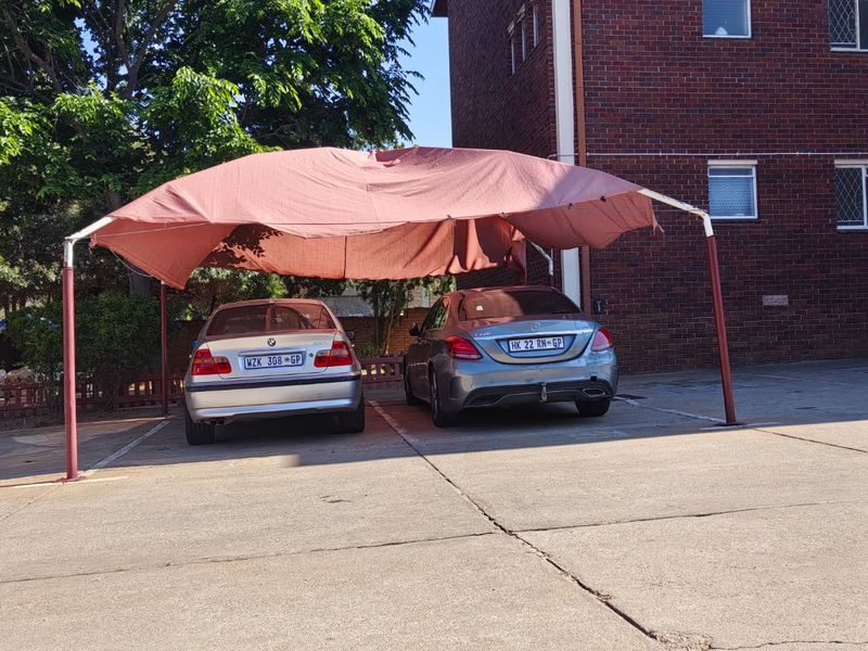 Outdoor parking area featuring a shade cloth carport structure situated on a concrete driveway adjacent to the brick apartment building.