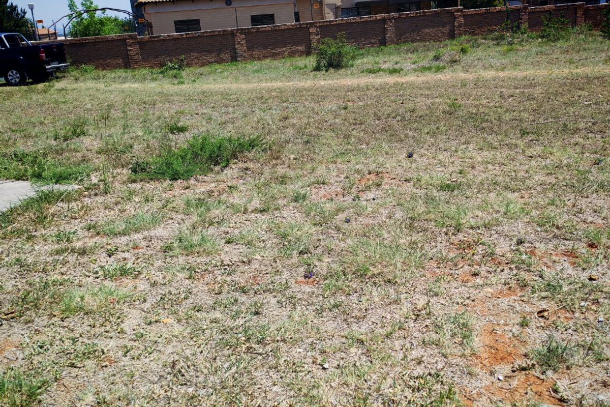 A vacant residential plot featuring natural grass cover, enclosed by a brick boundary wall with neighboring residential structures visible in the background.