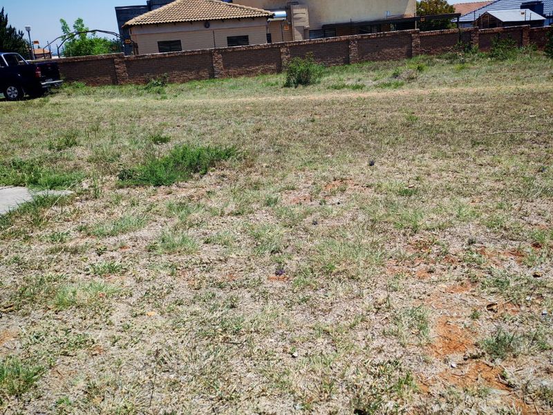 A vacant residential plot featuring natural grass cover, enclosed by a brick boundary wall with neighboring residential structures visible in the background.