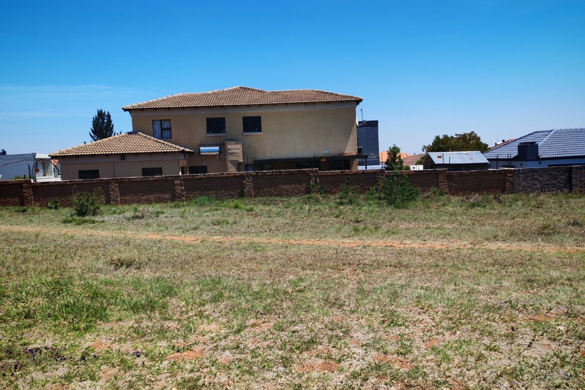 A vacant residential plot of land featuring a brick boundary wall with neighboring residential properties visible in the background.