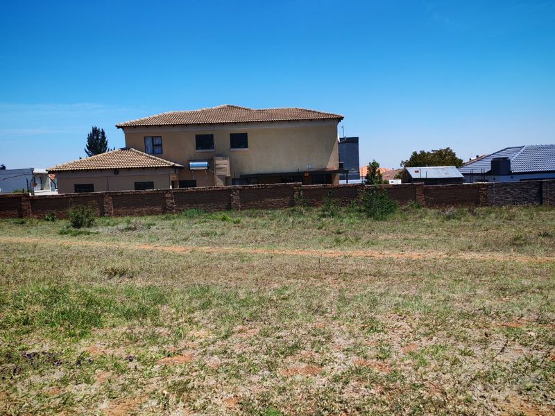 A vacant residential plot of land featuring a brick boundary wall with neighboring residential properties visible in the background.