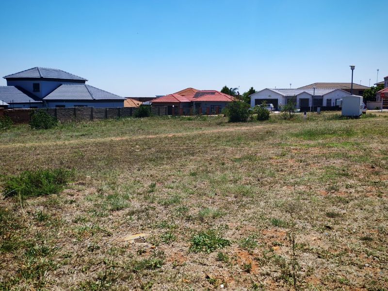 A vacant residential plot of land situated in a developed neighborhood, featuring natural ground cover and boundary walling.