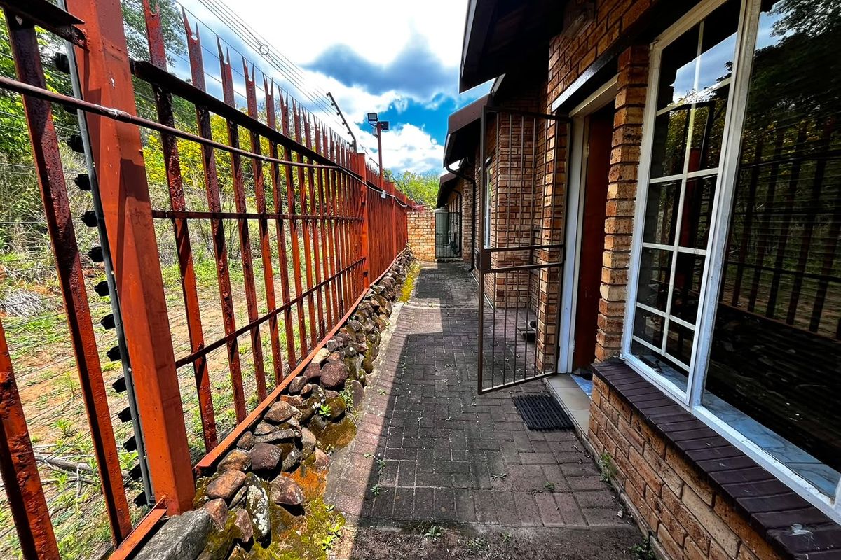 A narrow paved side walkway along a brick townhouse exterior, featuring a metal security fence with electric wiring and a stone retaining wall.