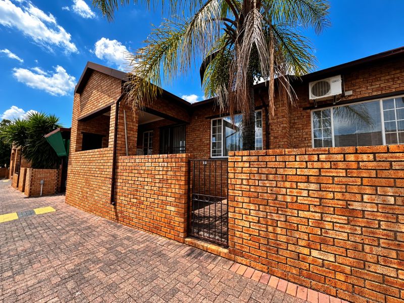 Front elevation of a face brick townhouse featuring a paved driveway, secure gated entrance, and exterior air conditioning unit.
