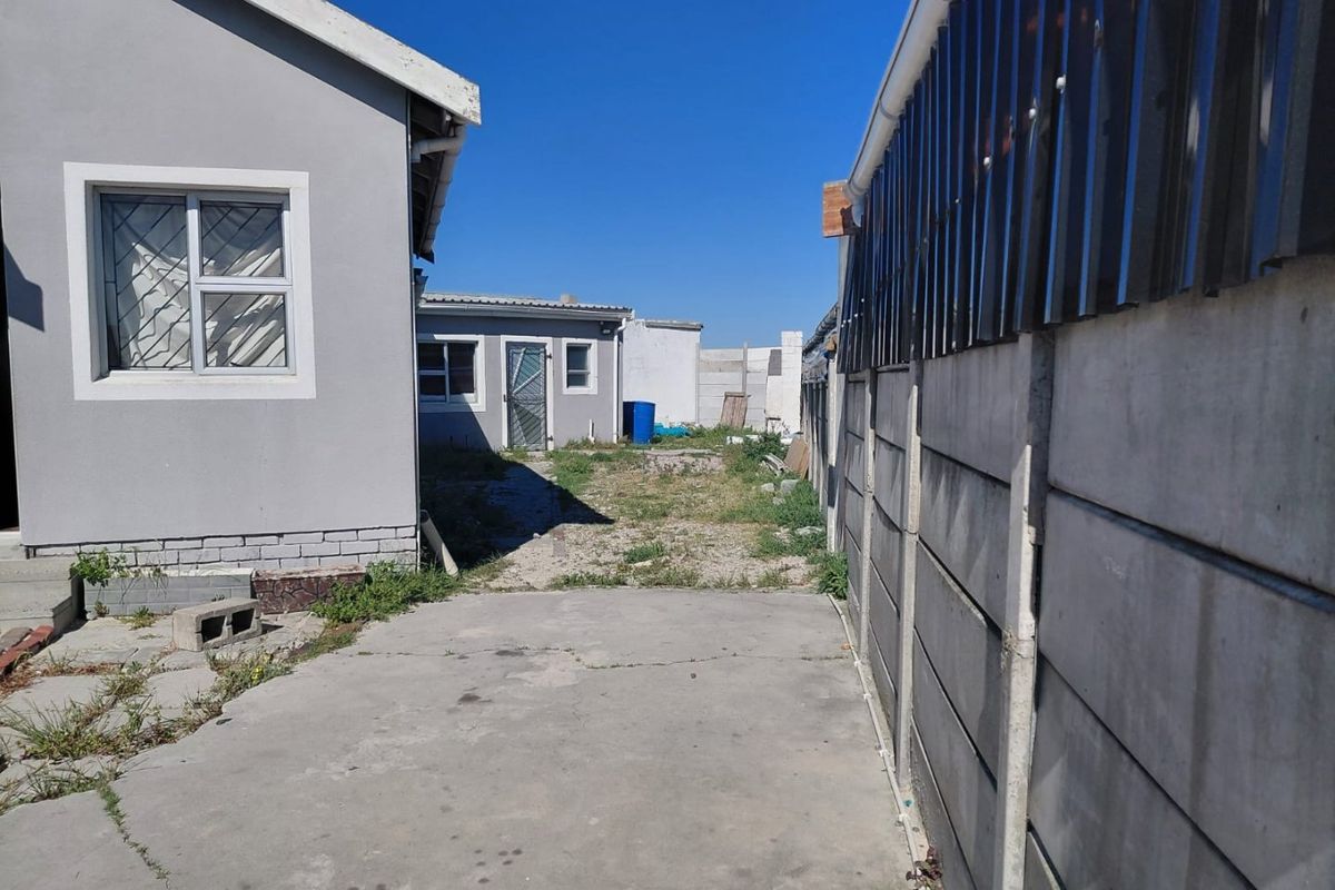 A rear exterior view showing a concrete driveway, precast boundary wall, and an unmaintained yard area between the main house and outbuilding.
