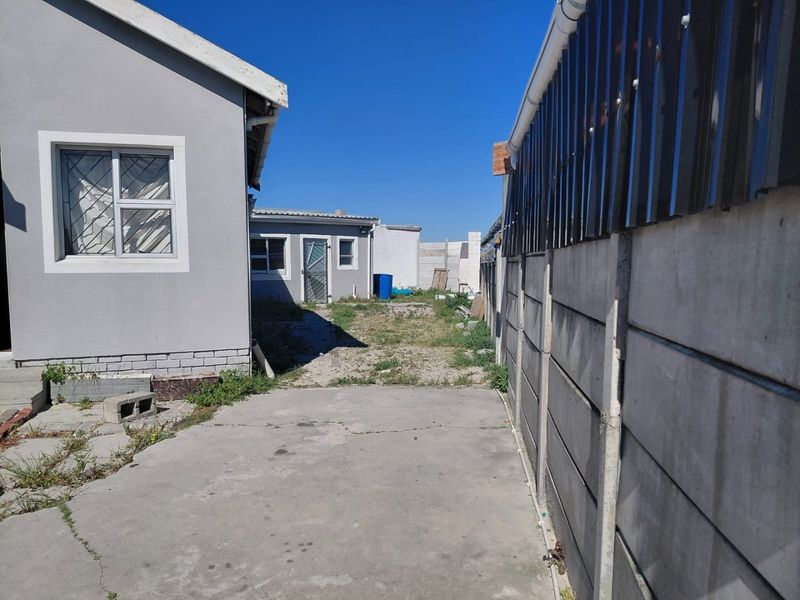 A rear exterior view showing a concrete driveway, precast boundary wall, and an unmaintained yard area between the main house and outbuilding.
