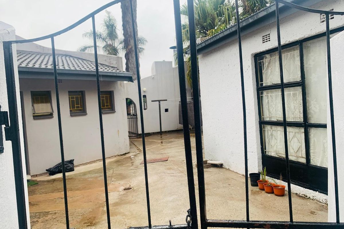 A view through a wrought iron gate into a paved rear courtyard area with white exterior walls and security-barred windows.