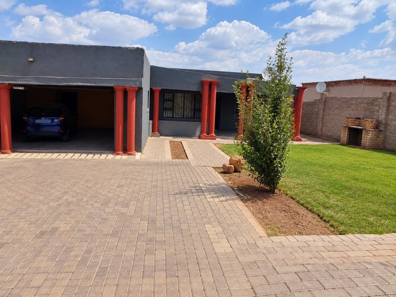 Front exterior view featuring a paved driveway, covered garage, decorative columns, and a garden area with a built-in brick braai.