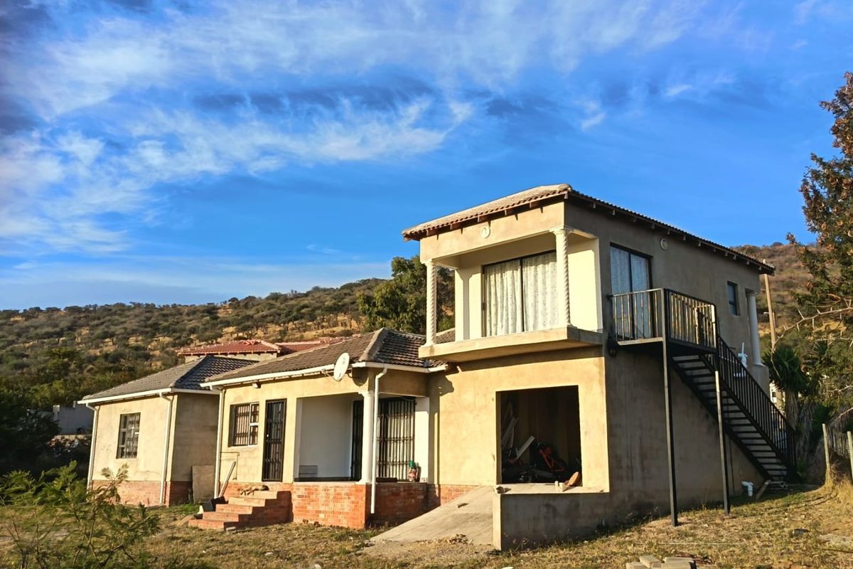 Rear view of a multi-level house featuring an external staircase, balcony, and unfinished exterior plaster work, set against a hillside landscape.