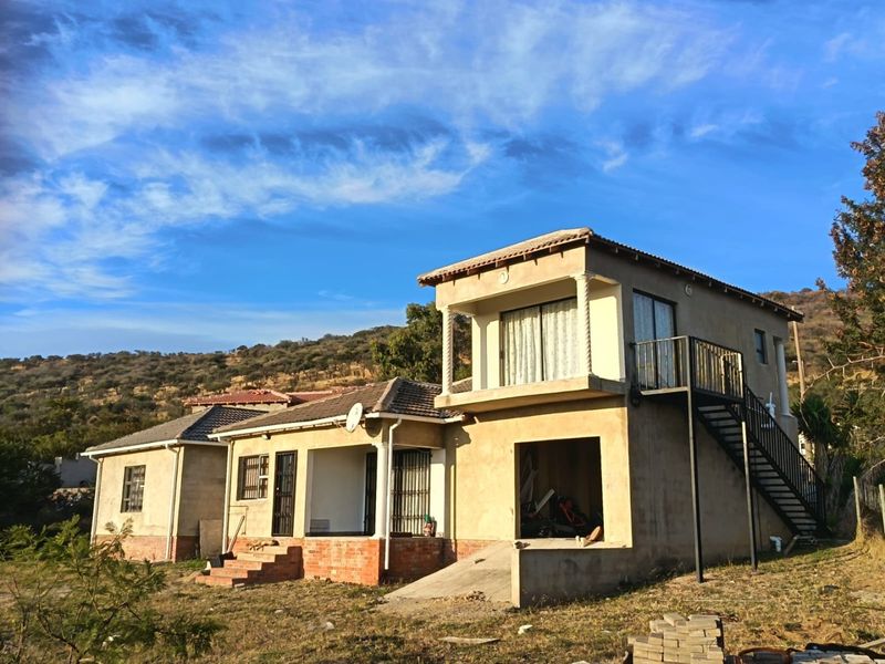 Rear view of a multi-level house featuring an external staircase, balcony, and unfinished exterior plaster work, set against a hillside landscape.