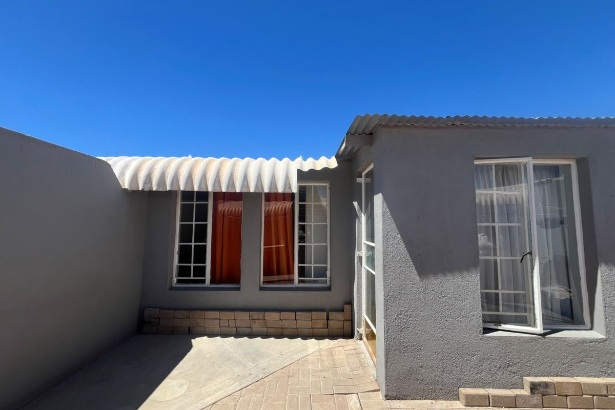 A rear exterior view of the property featuring a paved patio area, plastered walls, and a corrugated roof awning over the windows.