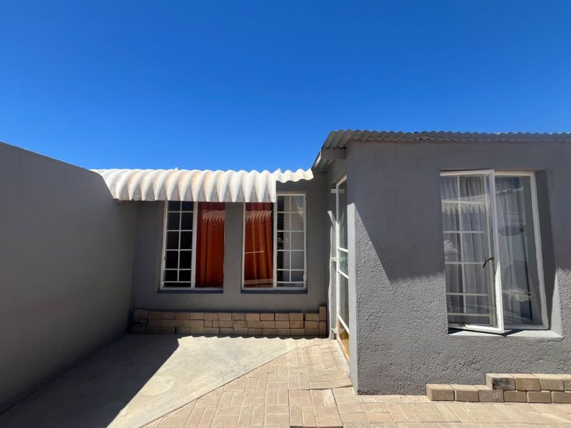 A rear exterior view of the property featuring a paved patio area, plastered walls, and a corrugated roof awning over the windows.
