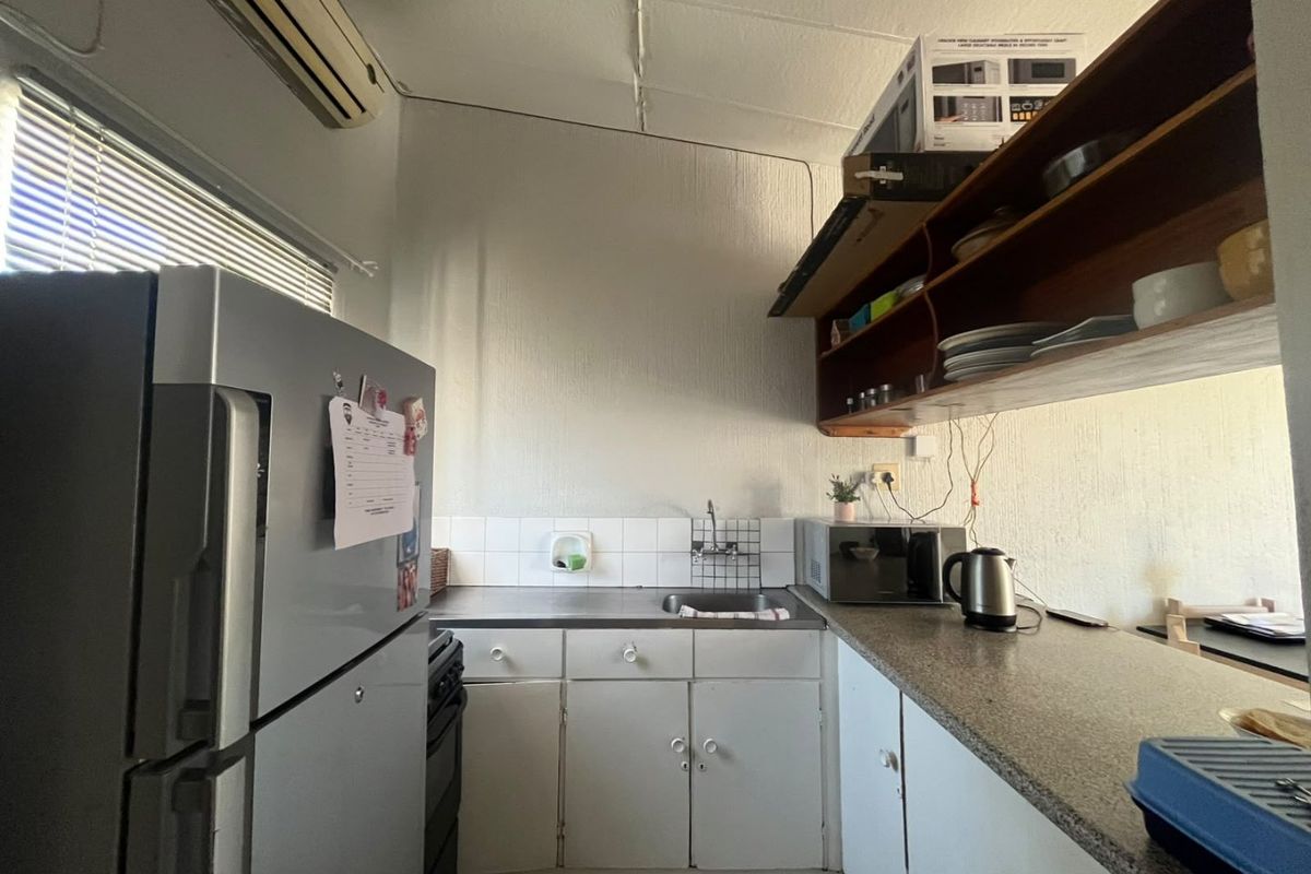 A compact kitchen featuring built-in cabinetry, a stainless steel sink, tiled backsplash, and wall-mounted shelving, providing a functional but dated preparation space.