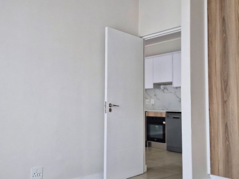 A clean, modern bedroom featuring built-in wood-finish cupboards and a view through to the adjacent kitchen area.