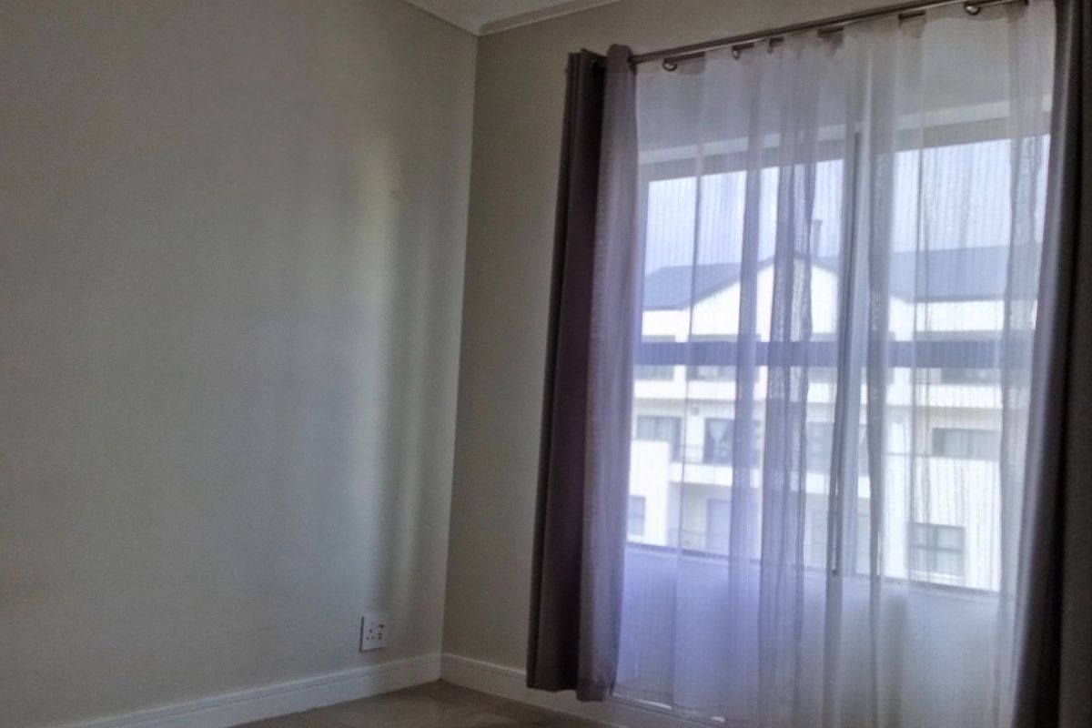 A bright, empty bedroom featuring neutral walls, carpeted flooring, and a window with installed curtain tracks.