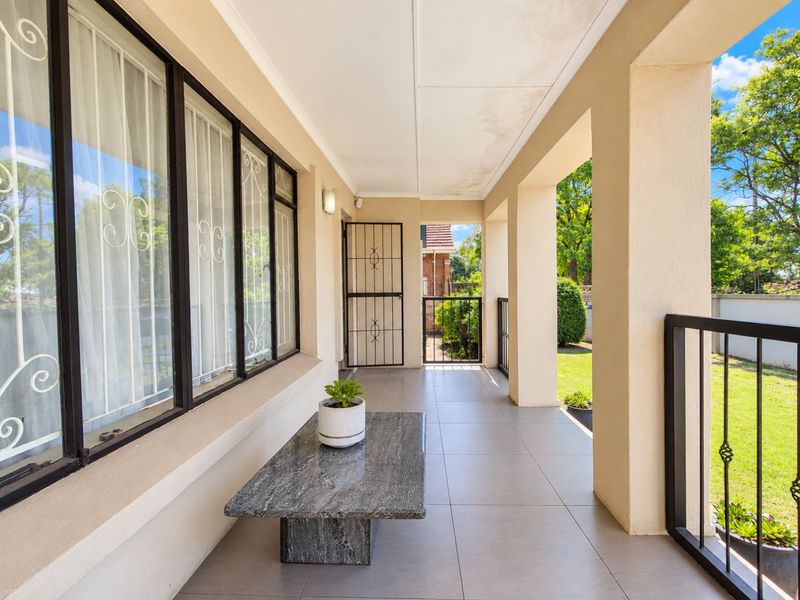 A covered patio area featuring tiled flooring, security gates, and structural pillars, overlooking a garden space.