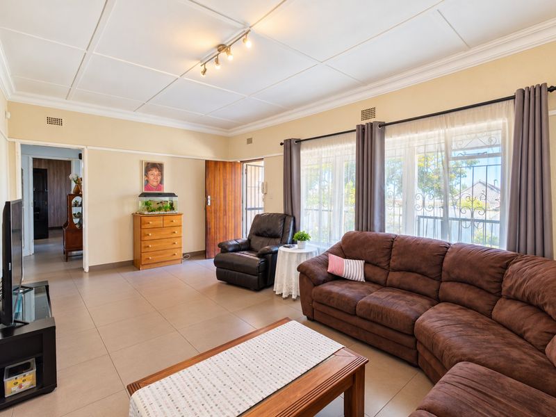 A spacious living room featuring tiled flooring, ceiling tiles, and large windows with security bars, offering a functional and well-lit living space.