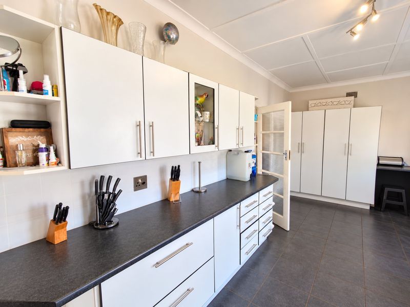 A functional scullery or utility area featuring extensive white built-in cabinetry, dark laminate countertops, tiled flooring, and a glass-paneled door.