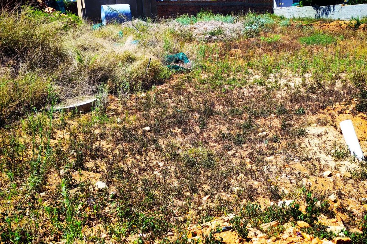 A vacant residential plot featuring uneven terrain and overgrown vegetation, situated in a developed neighborhood with surrounding modern residential properties.