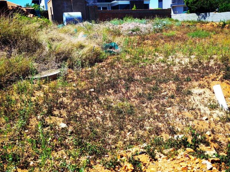 A vacant residential plot featuring uneven terrain and overgrown vegetation, situated in a developed neighborhood with surrounding modern residential properties.