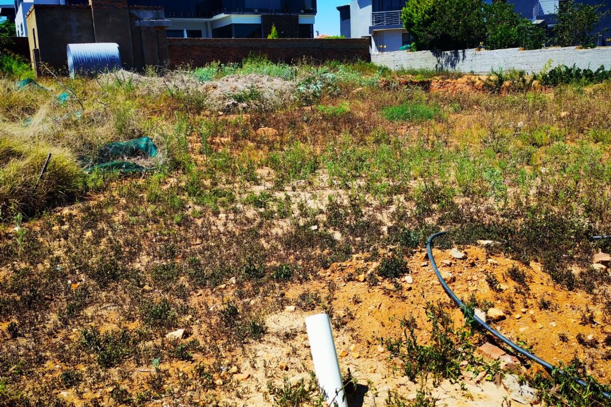 Vacant residential land plot featuring uneven terrain, overgrown vegetation, and existing boundary walls with nearby residential development.