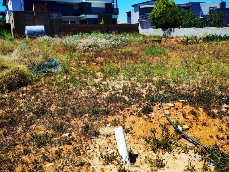 Vacant residential land plot featuring uneven terrain, overgrown vegetation, and existing boundary walls with nearby residential development.