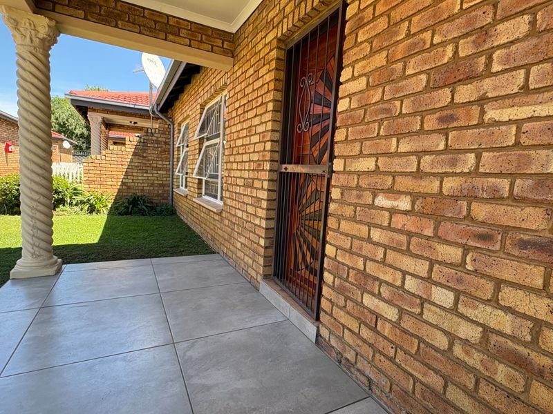 A covered patio area featuring face brick walls, tiled flooring, and a decorative column, providing access to the home through a security gate.
