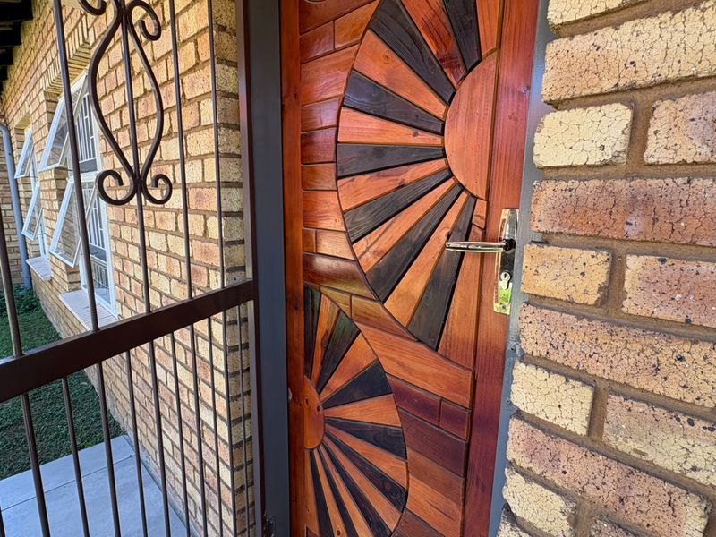 A close-up view of a decorative wooden front door set within a face brick exterior wall, featuring a security gate and aluminium window frames.
