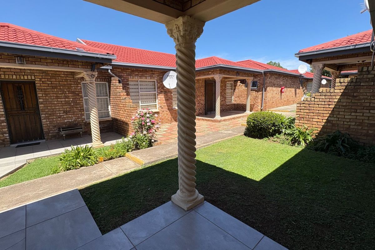 A well-maintained rear courtyard featuring brick exterior walls, a tiled roof, decorative spiral columns, and a paved patio area overlooking a small lawn.