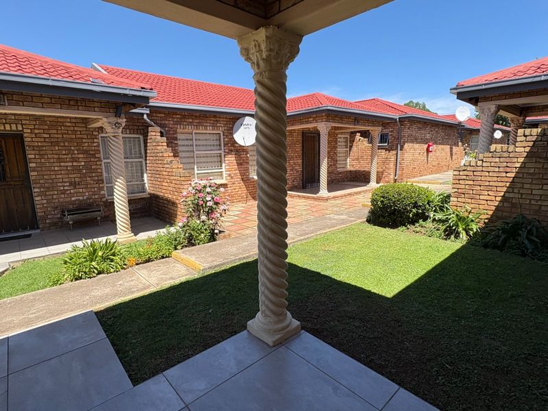 A well-maintained rear courtyard featuring brick exterior walls, a tiled roof, decorative spiral columns, and a paved patio area overlooking a small lawn.