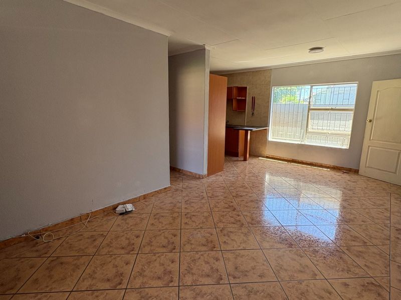 An open-plan living area featuring tiled flooring, neutral walls, and a compact kitchen corner with built-in cabinetry and security bars on the window.