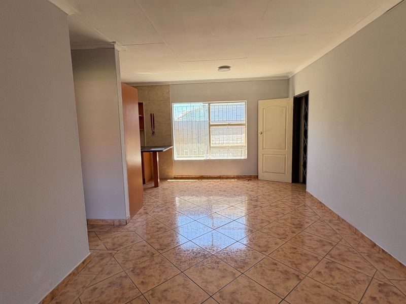 An open-plan living area featuring tiled flooring, neutral walls, and a window with security bars, offering a clean and versatile space.