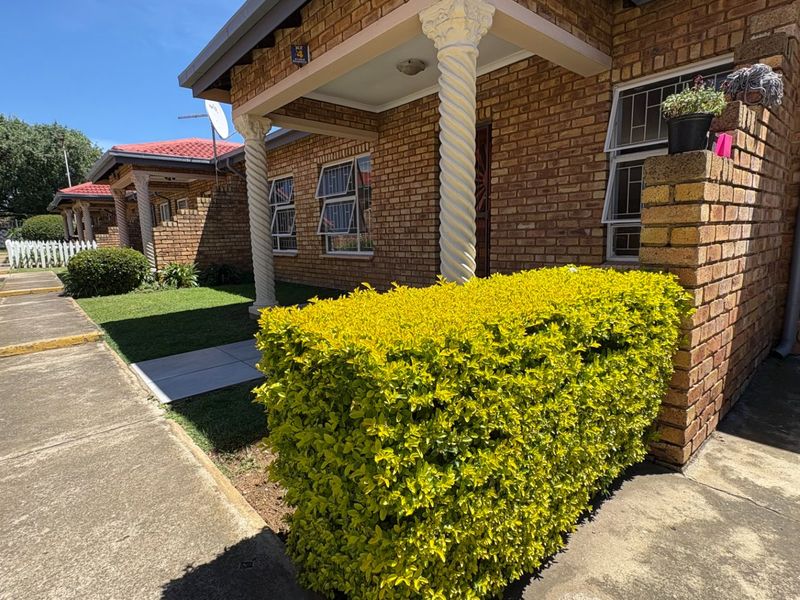 A well-maintained face brick residential unit featuring a covered porch with decorative columns, paved walkways, and established garden landscaping.