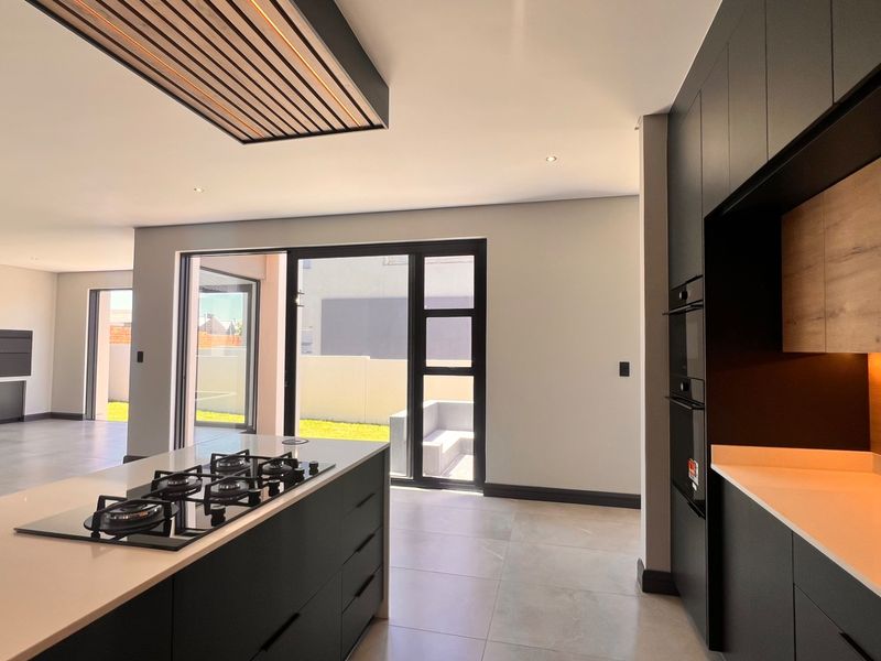 Modern open-plan kitchen featuring a central island with gas hob, sleek dark cabinetry, stone countertops, and seamless flow to the outdoor patio area.