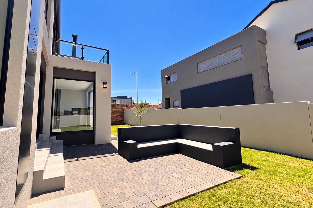 Modern rear exterior featuring a paved patio area with built-in masonry seating, a landscaped garden, and an upper-level balcony with glass balustrades.